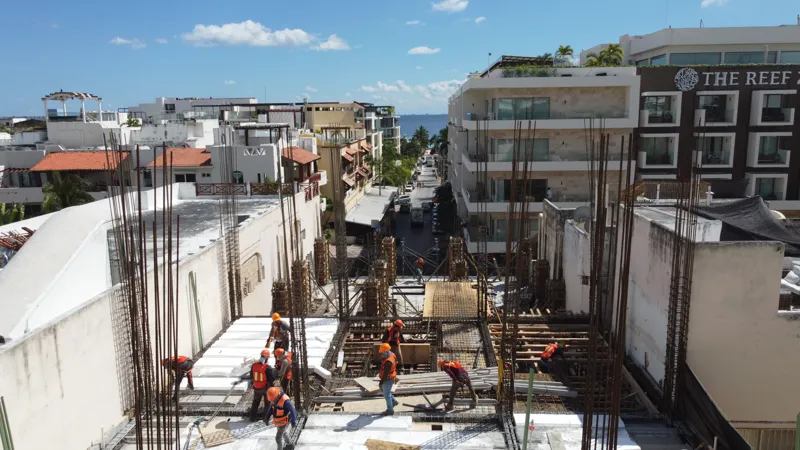 An aerial view of the Costera Mamitas construction site in Playa del Carmen, showing multiple workers in safety gear on a partially completed concrete slab, surrounded by wooden formwork for new columns and exposed rebar, with the vibrant blue Car...