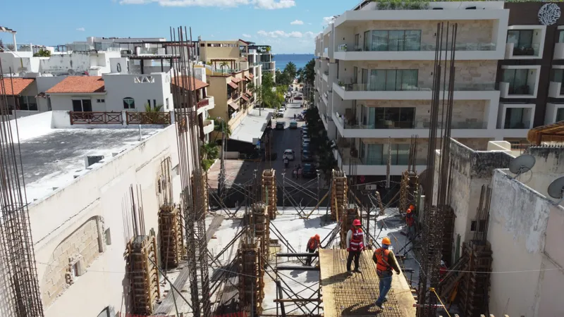 An aerial view of the Costera Mamitas construction site in Playa del Carmen, showing multiple workers in safety gear on a partially completed concrete slab, surrounded by wooden formwork for new columns and exposed rebar, with the vibrant blue Car...