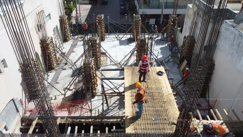 An aerial view of the Costera Mamitas construction site in Playa del Carmen, showing multiple workers in safety gear on a partially completed concrete slab, surrounded by wooden formwork for new columns and exposed rebar, with the vibrant blue Car...