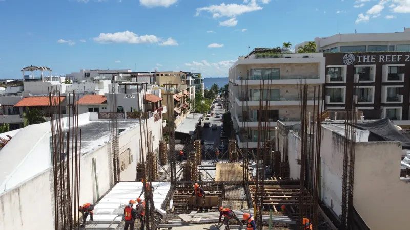 An aerial view of the Costera Mamitas construction site in Playa del Carmen, showing multiple workers in safety gear on a partially completed concrete slab, surrounded by wooden formwork for new columns and exposed rebar, with the vibrant blue Car...