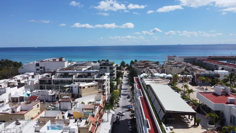 An aerial view of the Costera Mamitas construction site in Playa del Carmen, showing multiple workers in safety gear on a partially completed concrete slab, surrounded by wooden formwork for new columns and exposed rebar, with the vibrant blue Car...