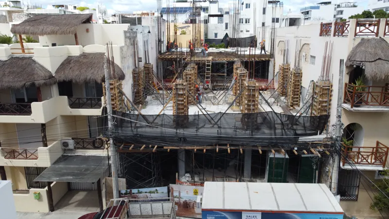 An aerial view of the Costera Mamitas construction site in Playa del Carmen, showing multiple workers in safety gear on a partially completed concrete slab, surrounded by wooden formwork for new columns and exposed rebar, with the vibrant blue Car...