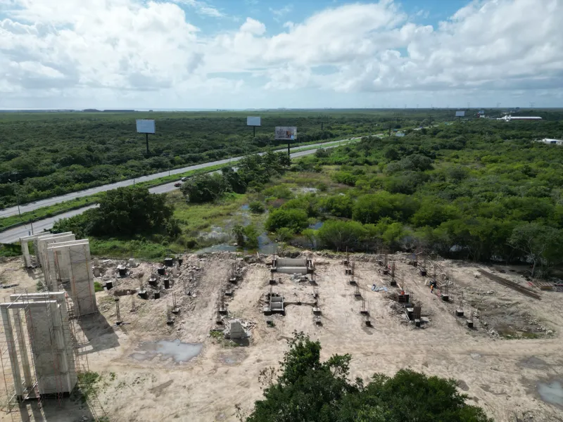 Aerial view of the Anthar development construction site in Playa del Carmen, showing multiple concrete columns and wall sections erected, alongside ongoing foundational work with rebar visible. A highway runs parallel to the site, with lush green ...