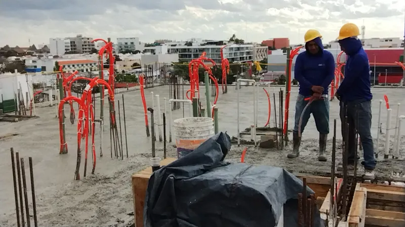 Construction workers in hard hats and safety vests are actively pouring and spreading concrete on an upper floor of the Ola de Mar development. The image shows rebar, orange electrical conduits, and fresh concrete, with other urban buildings visib...