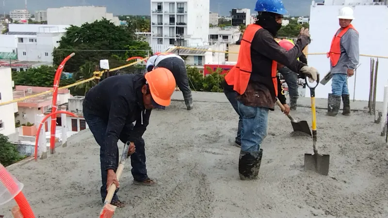 Construction workers in hard hats and safety vests are actively pouring and spreading concrete on an upper floor of the Ola de Mar development. The image shows rebar, orange electrical conduits, and fresh concrete, with other urban buildings visib...