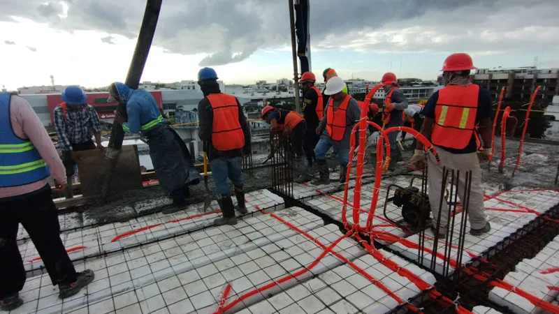 Construction workers in hard hats and safety vests are actively pouring and spreading concrete on an upper floor of the Ola de Mar development. The image shows rebar, orange electrical conduits, and fresh concrete, with other urban buildings visib...