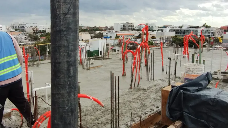 Construction workers in hard hats and safety vests are actively pouring and spreading concrete on an upper floor of the Ola de Mar development. The image shows rebar, orange electrical conduits, and fresh concrete, with other urban buildings visib...