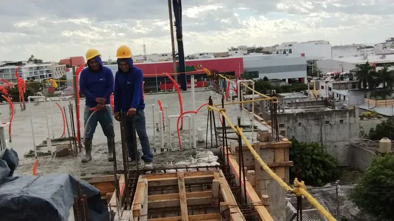 Construction workers in hard hats and safety vests are actively pouring and spreading concrete on an upper floor of the Ola de Mar development. The image shows rebar, orange electrical conduits, and fresh concrete, with other urban buildings visib...
