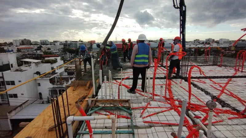 Construction workers in hard hats and safety vests are actively pouring and spreading concrete on an upper floor of the Ola de Mar development. The image shows rebar, orange electrical conduits, and fresh concrete, with other urban buildings visib...