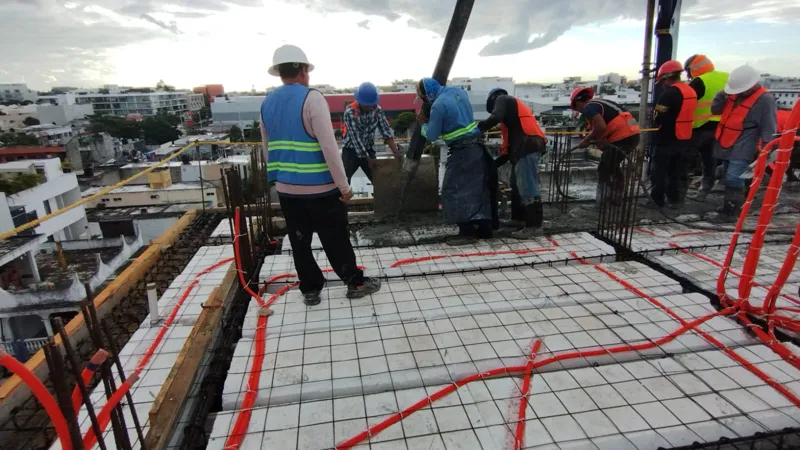 Construction workers in hard hats and safety vests are actively pouring and spreading concrete on an upper floor of the Ola de Mar development. The image shows rebar, orange electrical conduits, and fresh concrete, with other urban buildings visib...