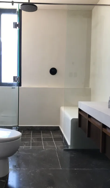 A modern bathroom vanity in a Humana studio unit, featuring a unique rough-hewn grey stone vessel sink with a black faucet, set on a white countertop with dark wood drawers, and a wooden-framed mirror above, reflecting a glass shower enclosure.