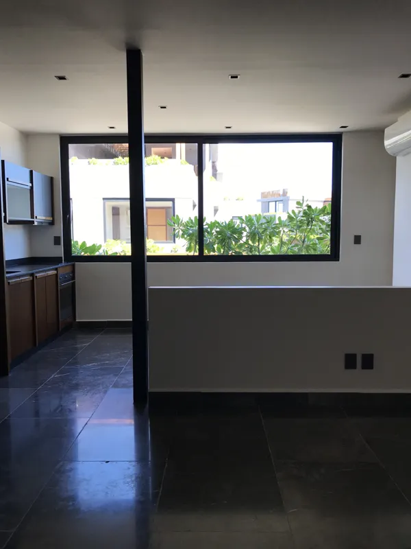 A modern bathroom vanity in a Humana studio unit, featuring a unique rough-hewn grey stone vessel sink with a black faucet, set on a white countertop with dark wood drawers, and a wooden-framed mirror above, reflecting a glass shower enclosure.