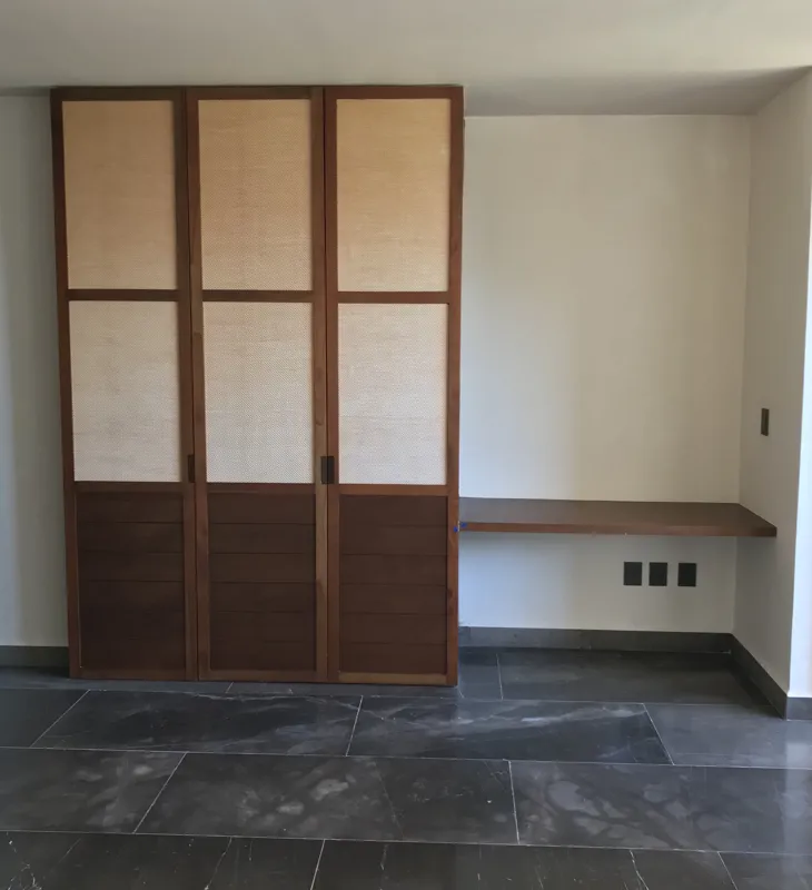 A modern bathroom vanity in a Humana studio unit, featuring a unique rough-hewn grey stone vessel sink with a black faucet, set on a white countertop with dark wood drawers, and a wooden-framed mirror above, reflecting a glass shower enclosure.