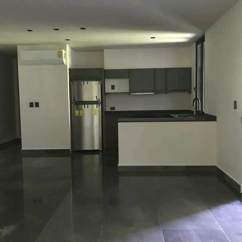 A modern kitchen in a Humana model unit, featuring dark grey floor tiles, white walls, grey upper and dark wood lower kitchen cabinets, a stainless steel refrigerator, a black countertop island, and a large window providing natural light.