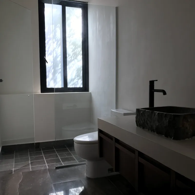 A modern kitchen in a Humana model unit, featuring dark grey floor tiles, white walls, grey upper and dark wood lower kitchen cabinets, a stainless steel refrigerator, a black countertop island, and a large window providing natural light.