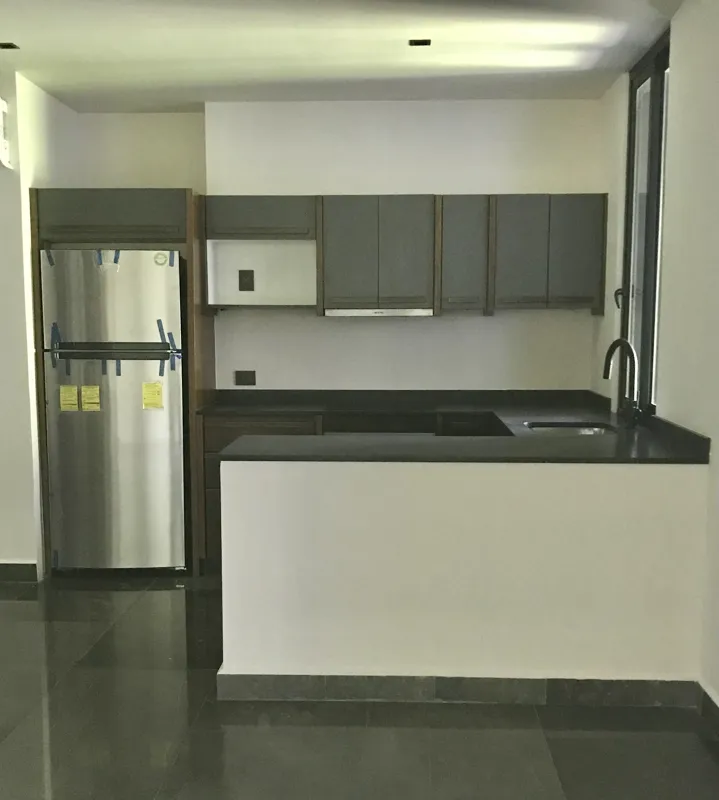 A modern kitchen in a Humana model unit, featuring dark grey floor tiles, white walls, grey upper and dark wood lower kitchen cabinets, a stainless steel refrigerator, a black countertop island, and a large window providing natural light.