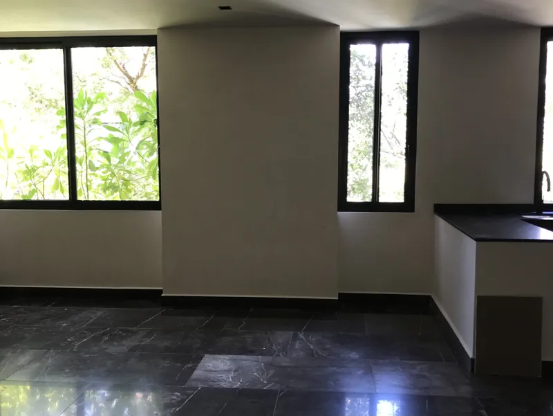A modern kitchen in a Humana model unit featuring dark cabinetry, black countertops, a stainless steel refrigerator, and dark tiled floors, with large windows offering views of lush greenery.