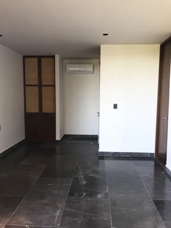 Interior view of a finished residential unit at Humana Tulum, featuring dark polished marble floors, white walls, a built-in wooden closet with sliding doors, an air conditioning unit, and a modern bathroom with a unique stone basin, black fixture...