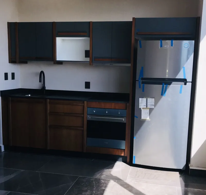 A modern kitchen in Humana's Unit 303D, featuring dark grey upper cabinets, wooden lower cabinets, a black sink, an integrated oven, and a stainless steel refrigerator with protective tape, all set against dark tiled flooring.
