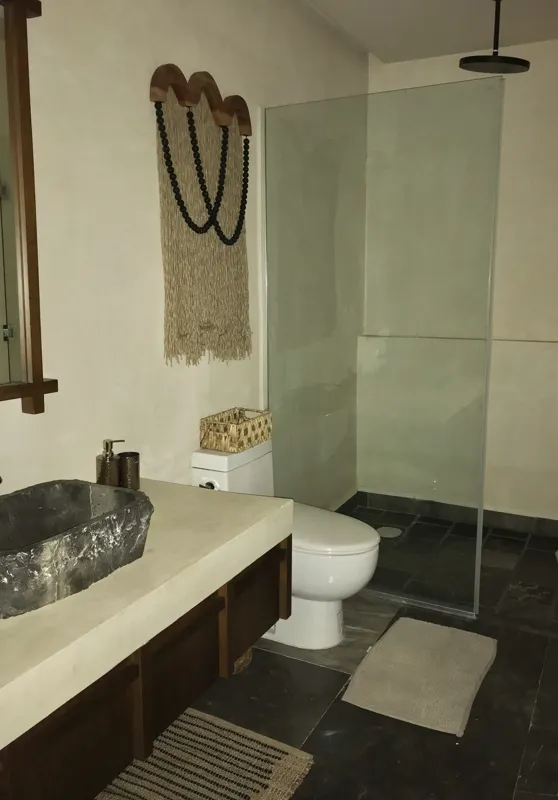 A modern kitchen in a Humana model unit, featuring dark cabinetry, stainless steel appliances, a black sink, a light wood breakfast bar with two woven stools, and decorative wall plates, illuminated by natural light from a window.