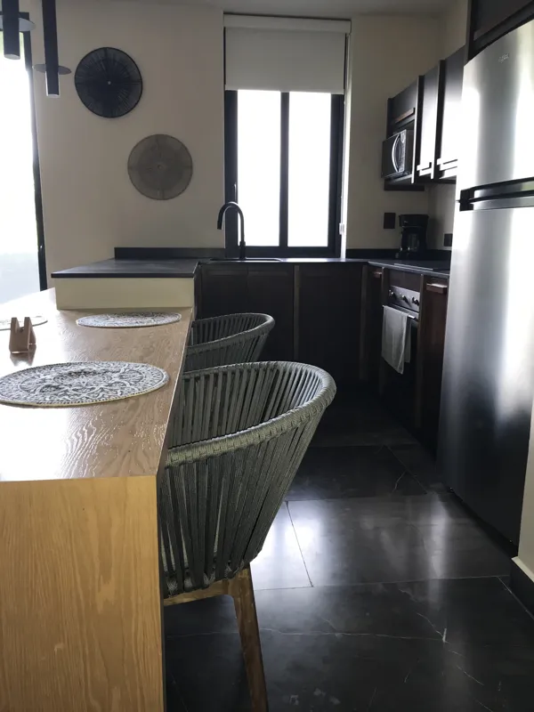 A modern kitchen in a Humana model unit, featuring dark cabinetry, stainless steel appliances, a black sink, a light wood breakfast bar with two woven stools, and decorative wall plates, illuminated by natural light from a window.