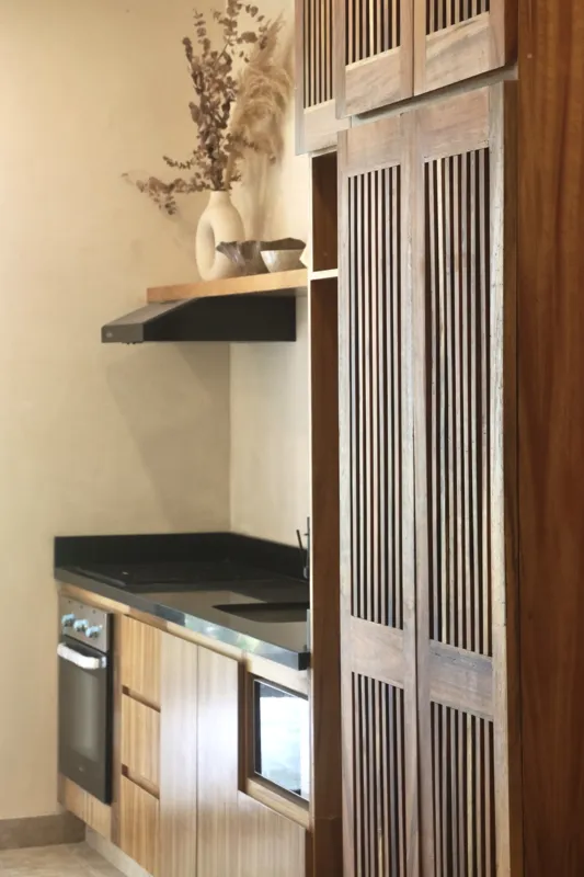 A modern kitchen in an Uxmal unit featuring warm wooden cabinetry, a black countertop with an integrated sink, a built-in oven, microwave, and a round wooden dining table with chairs, all under soft recessed lighting.