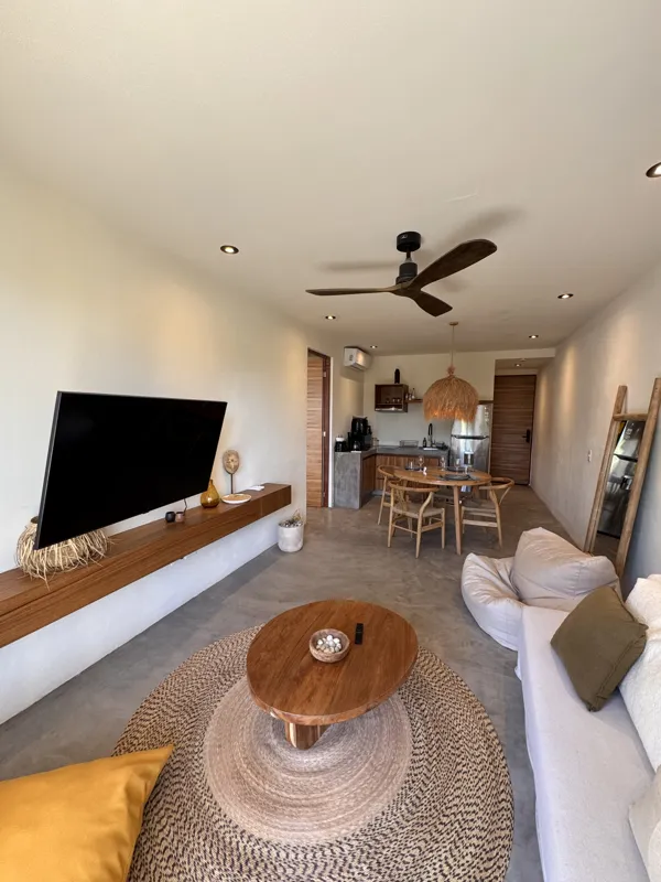 A wide-angle view of a modern apartment's dining area and kitchen in Gold Garden, featuring a round wooden dining table with four chairs, a large woven straw pendant light, and a compact kitchen with concrete countertops and stainless steel applia...