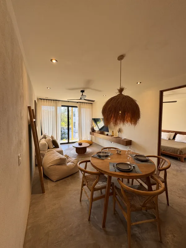 A wide-angle view of a modern apartment's dining area and kitchen in Gold Garden, featuring a round wooden dining table with four chairs, a large woven straw pendant light, and a compact kitchen with concrete countertops and stainless steel applia...