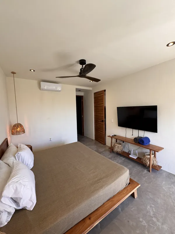 A wide-angle view of a modern apartment's dining area and kitchen in Gold Garden, featuring a round wooden dining table with four chairs, a large woven straw pendant light, and a compact kitchen with concrete countertops and stainless steel applia...