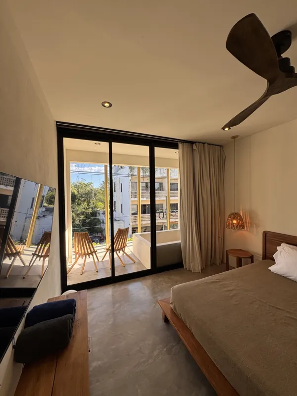 A wide-angle view of a modern apartment's dining area and kitchen in Gold Garden, featuring a round wooden dining table with four chairs, a large woven straw pendant light, and a compact kitchen with concrete countertops and stainless steel applia...
