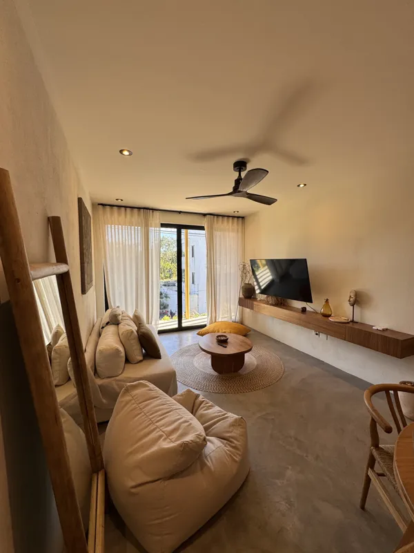 A wide-angle view of a modern apartment's dining area and kitchen in Gold Garden, featuring a round wooden dining table with four chairs, a large woven straw pendant light, and a compact kitchen with concrete countertops and stainless steel applia...