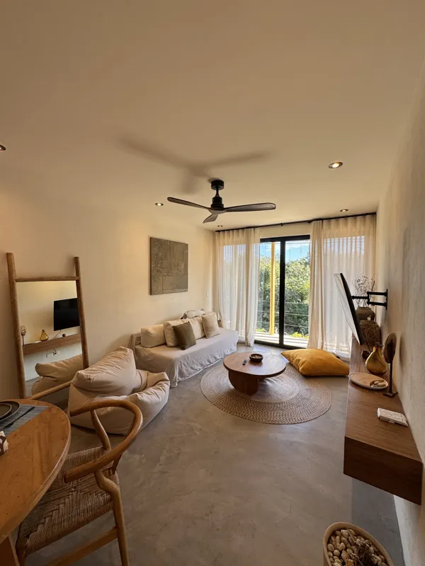 A wide-angle view of a modern apartment's dining area and kitchen in Gold Garden, featuring a round wooden dining table with four chairs, a large woven straw pendant light, and a compact kitchen with concrete countertops and stainless steel applia...