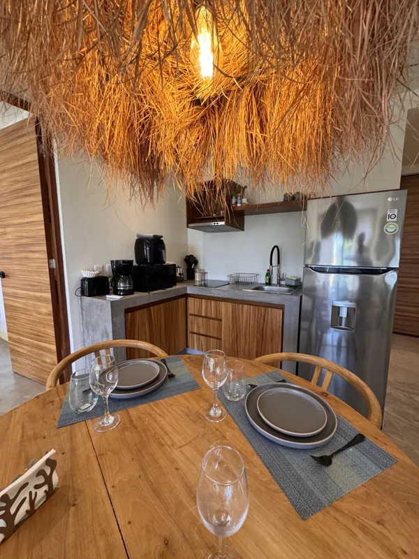 A wide-angle view of a modern apartment's dining area and kitchen in Gold Garden, featuring a round wooden dining table with four chairs, a large woven straw pendant light, and a compact kitchen with concrete countertops and stainless steel applia...