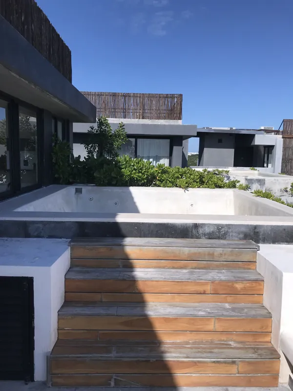 A luxurious rooftop terrace at Humana featuring a built-in jacuzzi with wooden steps, surrounded by modern architecture and lush greenery under a clear blue sky, showcasing finished outdoor amenities.