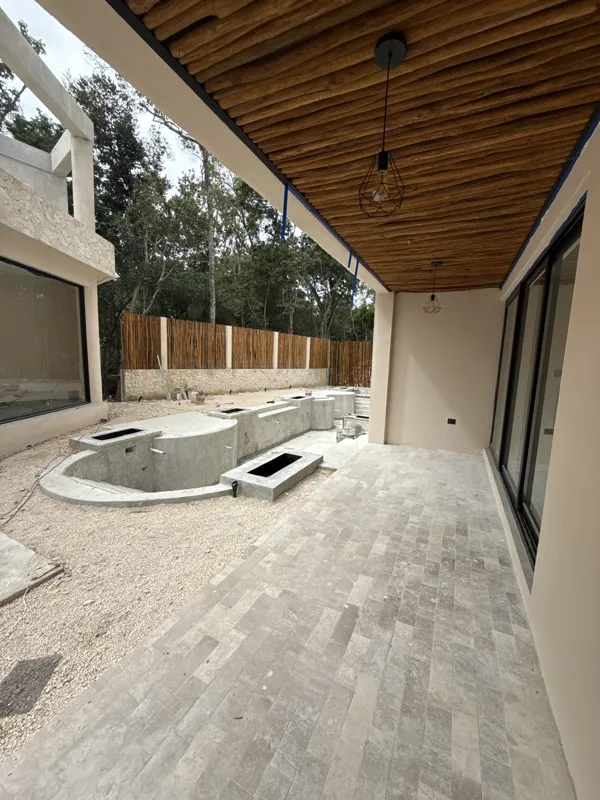 A wide shot of a garden unit's outdoor patio at Itza Selva, featuring light grey rectangular floor tiles, a natural wood log ceiling with two modern black pendant lights, and a concrete pool/water feature structure under construction. A gravel are...