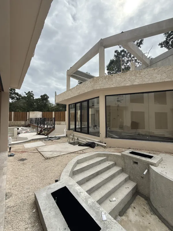 Detailed visual update of Itza Selva's common areas and building exterior. The images show significant construction progress, including the installation of large black-framed glass sliding doors on the main building, ongoing landscaping with grave...