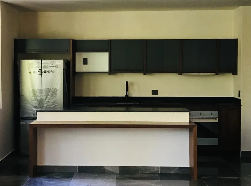 A modern kitchen in a Humana unit, featuring dark cabinetry, a central island with a light countertop, a stainless steel refrigerator still wrapped in protective plastic, and an integrated oven, all set against light-colored walls and dark floor t...