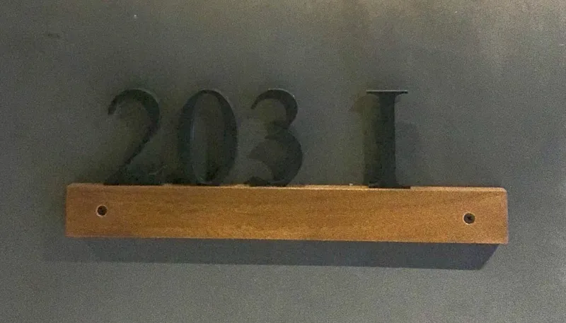A modern kitchen in a Humana unit, featuring dark cabinetry, a central island with a light countertop, a stainless steel refrigerator still wrapped in protective plastic, and an integrated oven, all set against light-colored walls and dark floor t...