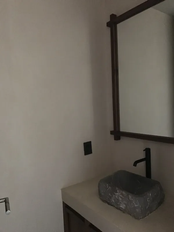 A modern bathroom vanity featuring a dark stone vessel sink, a black faucet, and a rectangular mirror with a dark wooden frame, set above a light-colored countertop and dark wood cabinetry.