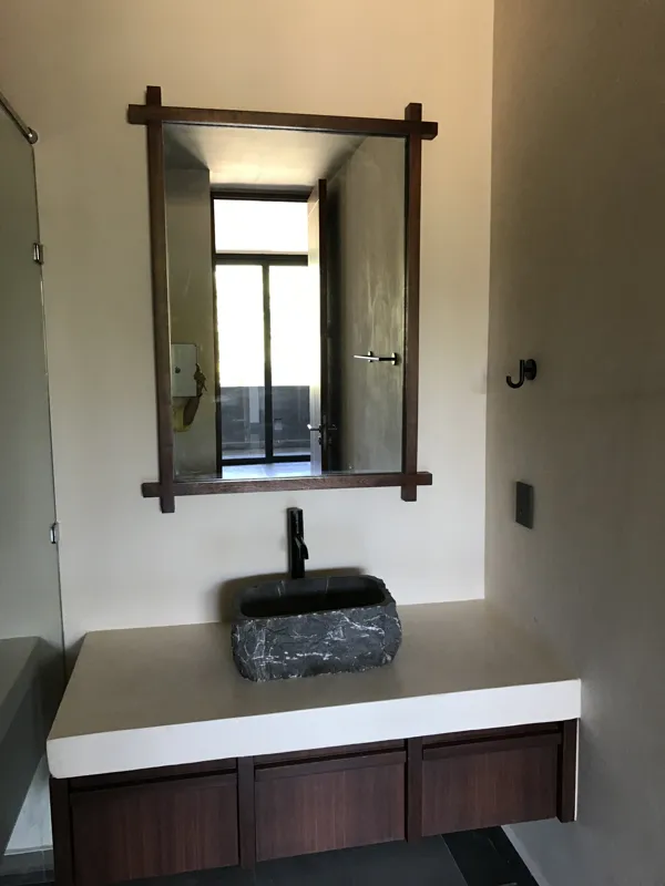 A modern bathroom vanity featuring a dark stone vessel sink, a black faucet, and a rectangular mirror with a dark wooden frame, set above a light-colored countertop and dark wood cabinetry.
