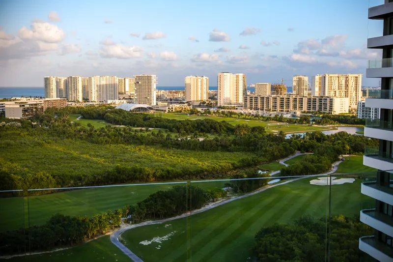 A beautifully furnished living room and balcony in a Woha residence, featuring modern decor, comfortable seating, and a coffee table with pastries, overlooking the Puerto Cancún skyline, a lush golf course, and distant natural reserve.