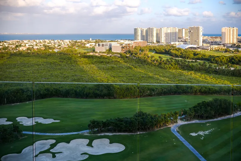 A beautifully furnished living room and balcony in a Woha residence, featuring modern decor, comfortable seating, and a coffee table with pastries, overlooking the Puerto Cancún skyline, a lush golf course, and distant natural reserve.