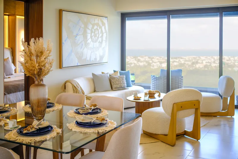 A beautifully furnished living room and balcony in a Woha residence, featuring modern decor, comfortable seating, and a coffee table with pastries, overlooking the Puerto Cancún skyline, a lush golf course, and distant natural reserve.