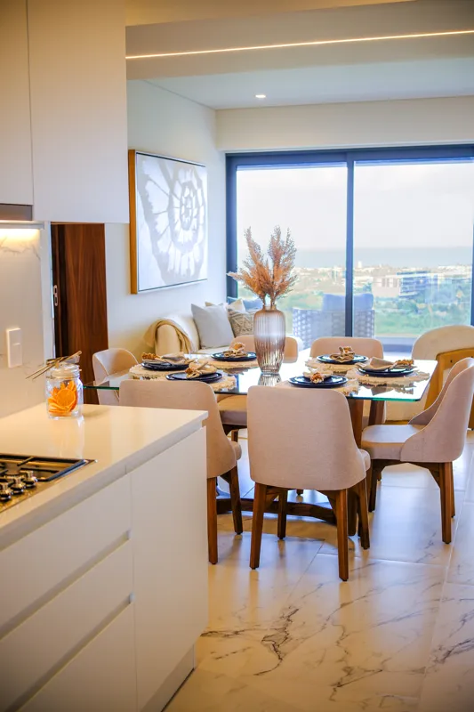 A beautifully furnished living room and balcony in a Woha residence, featuring modern decor, comfortable seating, and a coffee table with pastries, overlooking the Puerto Cancún skyline, a lush golf course, and distant natural reserve.