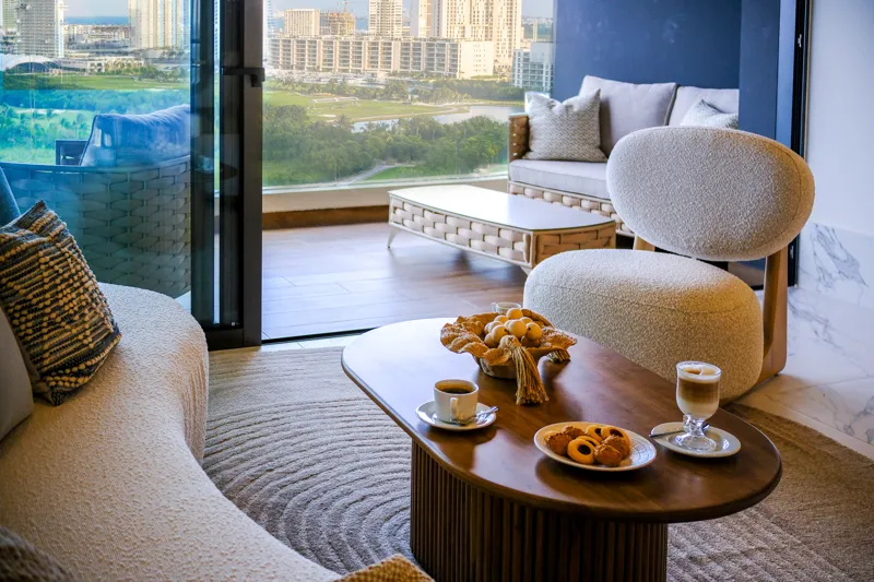 A beautifully furnished living room and balcony in a Woha residence, featuring modern decor, comfortable seating, and a coffee table with pastries, overlooking the Puerto Cancún skyline, a lush golf course, and distant natural reserve.