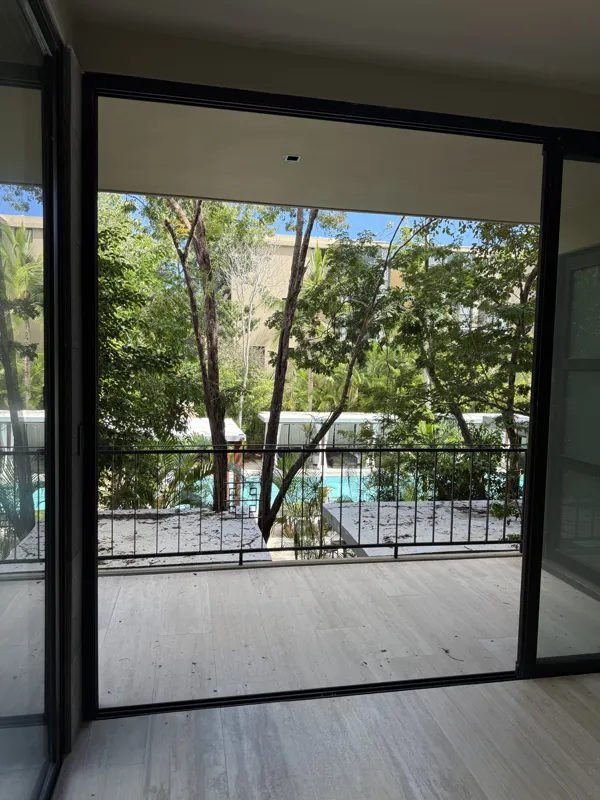 A bright, empty living room in an Arthouse Signature apartment, featuring light-colored tiled floors, an installed air conditioning unit, and expansive sliding glass doors leading to a private balcony with views of a lush tropical landscape and a ...