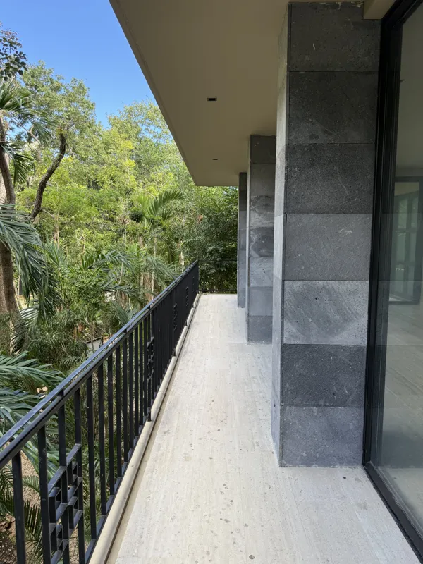 A bright, empty living room in an Arthouse Signature apartment, featuring light-colored tiled floors, an installed air conditioning unit, and expansive sliding glass doors leading to a private balcony with views of a lush tropical landscape and a ...