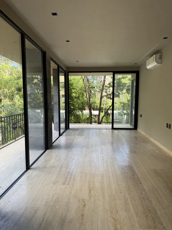 A bright, empty living room in an Arthouse Signature apartment, featuring light-colored tiled floors, an installed air conditioning unit, and expansive sliding glass doors leading to a private balcony with views of a lush tropical landscape and a ...