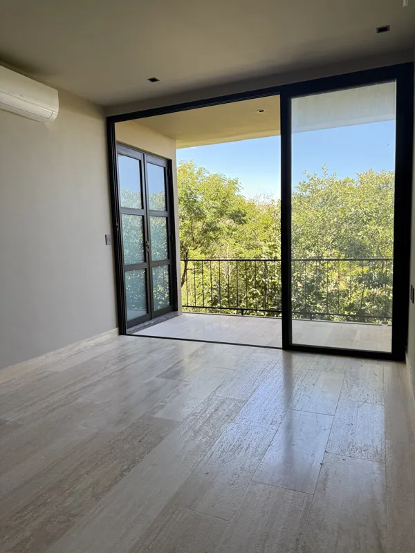 A bright, finished interior room in an Arthouse Signature one-bedroom apartment, featuring light-colored tiled floors, light grey walls, an installed air conditioning unit, and a large sliding glass door opening to a private balcony with lush gree...