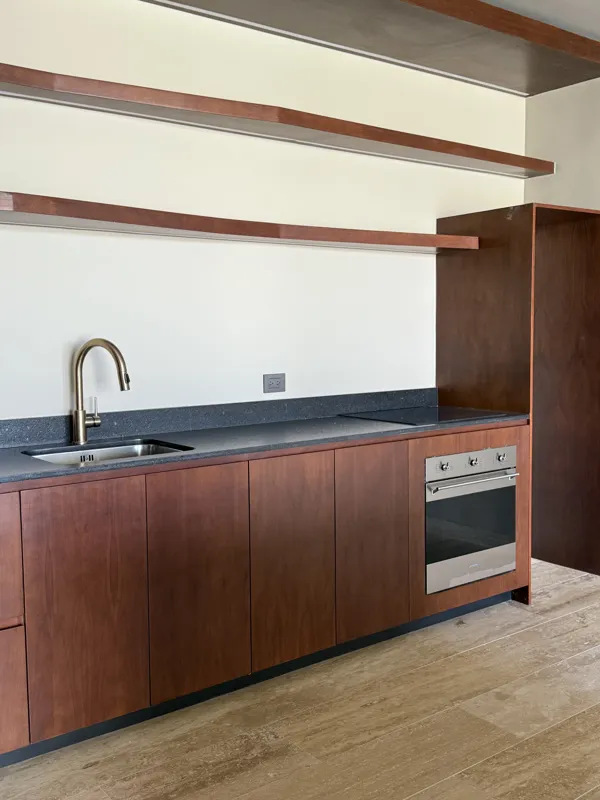 A bright, finished interior room in an Arthouse Signature one-bedroom apartment, featuring light-colored tiled floors, light grey walls, an installed air conditioning unit, and a large sliding glass door opening to a private balcony with lush gree...